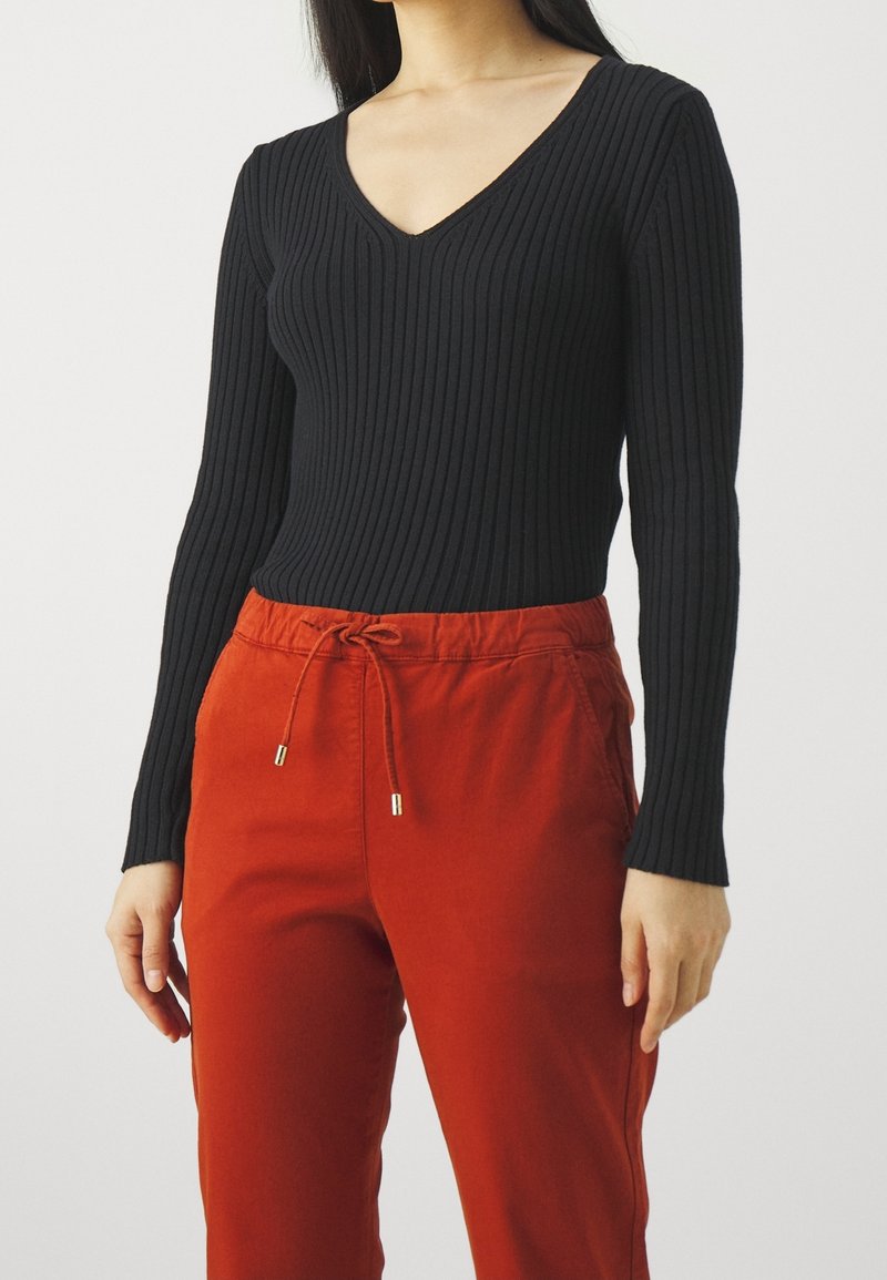 Femme portant un haut noir côtelé à manches longues avec col en V et un pantalon taille haute couleur rouille avec une taille à cordon.