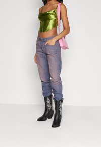 Top corto in satin verde con design drappeggiato, jeans blu sbiaditi e stivali da cowboy in pelle nera con ricami bianchi, abbinati a una borsa rosa.