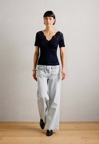 Námořnická blůza s krajkovým výstřihem, přiléhavý střih, krátké rukávy. Světle modré široké džíny. Černé kotníkové boty, na neutrálním pozadí.