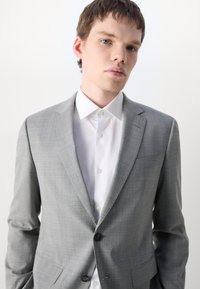 Giacca su misura grigia chiara con un sottile motivo a quadri, chiusura a un bottone, rever a forma di notched, abbinata a una camicia bianca con colletto.