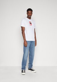 Camiseta blanca de algodón con un colorido estampado gráfico en el pecho, combinada con jeans azul claro y zapatillas negras con suelas blancas.