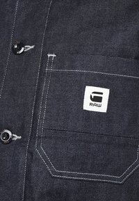 Tmavě modrá denimová košile se zapínáním na knoflíky, bílým šitím a čtvercovou nášivkou s logem na hrudní kapse. Hladká textura a strukturovaný střih.