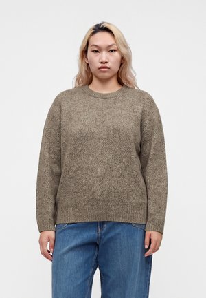 Ung person med langt blondt hår iført en brun strikket sweater og blå jeans, stående mod en ensartet lys baggrund.
