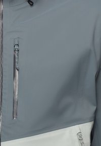 Close-up van een blauw-witte jas met twee verticale waterdichte zakken met rits, waarbij de textuur van de stof en details van de rits zichtbaar zijn.