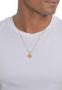 Ogrlica z zlato obeskom v obliki teksturiranega kroga, obešena na tanki verižici, nošena preko bele preproste majice.
