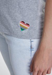 Chemise grise à manches courtes avec un patch en forme de cœur multicolore brodé à rayures. Portée avec un jean en denim léger. Texture en coton douce.