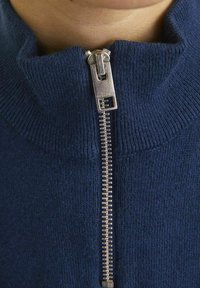 Un pull zippé tricoté bleu marine avec un col haut et des détails en zip argenté, offrant une surface texturée et des accents côtelés.