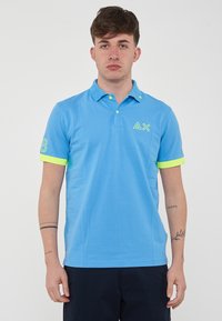 Polo di colore azzurro chiaro realizzato in cotone, con colletto, dettagli neon giallo sulle maniche e sui bottoni, e un logo grafico sul petto.