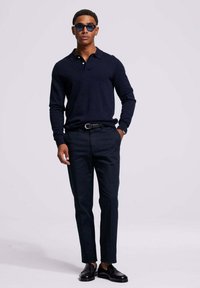 Marineblauwe longsleeve polo, slim-fit donkere broek, zwarte riem, glanzende zwarte schoenen en blauwe zonnebril, met een gladde textuur door het geheel.