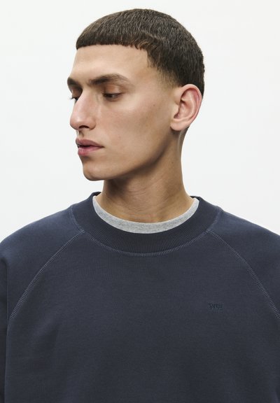 Mladi moški s kratkimi temnimi lasmi, oblečen v temno modro majico z okroglim izrezom čez sivo srajco, gleda levo, na preprostem belem ozadju.