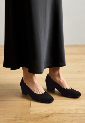 Cizme negre din suede cu un design clasic; vârf rotund și toc bloc; purtate cu o fustă neagră fluidă pe un podea din lemn.