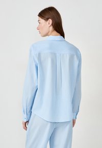 Mujer con cabello castaño que lleva una camisa de manga larga azul clara y pantalones a juego, de pie mirando hacia el fondo plano.
