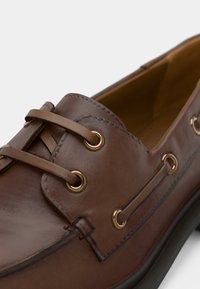 Κοντινή λήψη ενός καφέ δερμάτινου παπουτσιού σκάφους με δερμάτινες κορδόνες που περνάνε από χρυσά μάτια και λεπτομερή ραφή.