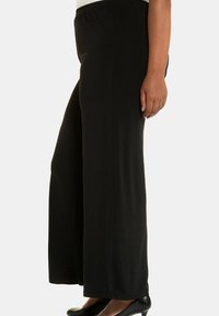 Kobieta w luźnych czarnych spodniach typu wide-leg, czarnych niskich obcasach i białej bluzce, pokazana od talii do stóp na jednolitym tle.