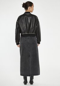 Giubbotto bomber in pelle nera con polsini a costine, abbinato a una lunga gonna di jeans scura e stivali neri fin sopra il ginocchio. Texture liscia, design casual.