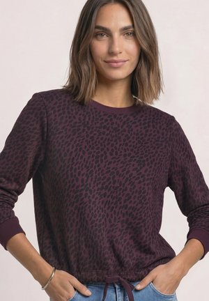Kvinde med skulderlangt brunt hår iført en mørkelilla leopardmønstret bluse med lange ærmer og lyseblå jeans, hænderne i lommerne.