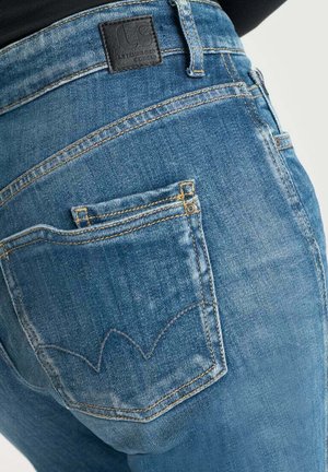 Les jeans en denim bleu présentent un motif de couture courbé distinctif sur les poches arrière et une étiquette en cuir texturée au niveau de la taille.