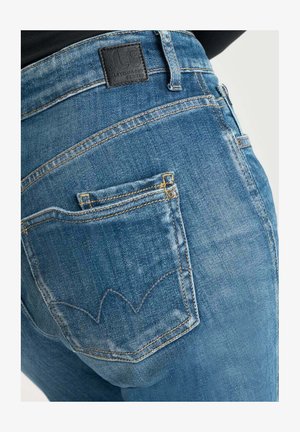 Blue denim jeans feature a distinctive curved stitching pattern on the back pockets and a textured leather tag at the waistband.
