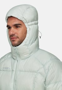 Man in een lichtgrijze pufferjack met opgetrokken capuchon, met de rits gesloten en het Mammut-logo zichtbaar op de borst, met het gezicht iets naar links gericht.