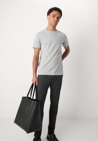 Camiseta gris de manga corta con un pequeño logo rojo, combinada con pantalones oscuros y sosteniendo una bolsa de tela negra con letras en relieve.