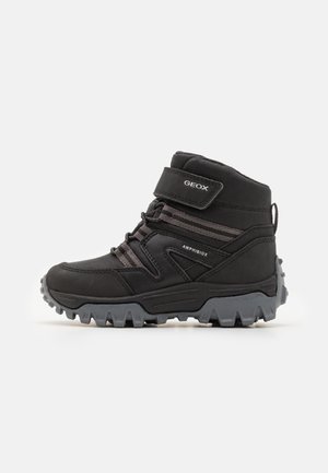 Bottes de cheville noires imperméables avec une semelle en caoutchouc, une sangle Velcro, des lacets élastiques et des accents en tissu texturé pour la stabilité et l'adhérence.