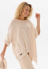 Jasno beżowy dzianinowy poncz z luźnym krojem, z podwiniętymi mankietami, akcentującym bocznym szwem i prążkowanym wykończeniem u dołu. Jasny kolor i miękka tekstura.