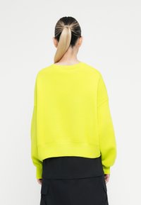 Neon rumena majica s kratkim krojem, spuštenimi rameni in rebrastimi manšeti, v kombinaciji s črnimi hlačami z stranskimi žepi.