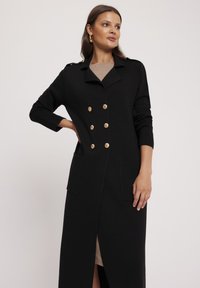 Czarny długi płaszcz z podwójnym zapięciem, złotymi guzikami, dwoma przednimi kieszeniami i długością do połowy uda, wykonany z teksturowanej tkaniny.