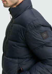 Tumši zils polsterēts jakas ar augstu apkakli, ar iešūtu priekšpusi un sānu kabatām, teksturēts audums un logo uz piedurknes.