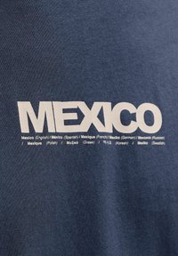 Σκούρο μπλε ύφασμα με τη λέξη «MEXICO» σε μεγάλα λευκά γράμματα και μεταφράσεις της λέξης «Μεξικό» σε πολλές γλώσσες από κάτω.
