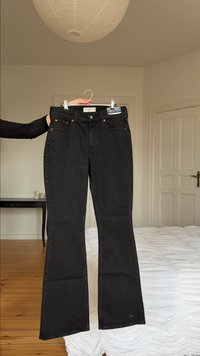 Άτομο που κρατάει μαύρο τζιν με ψηλή μέση και καμπάνα σε κρεμάστρα, μέσα σε υπνοδωμάτιο όπου είναι ορατά ένα κρεβάτι, μια πόρτα, ένα τραπέζι και ένα φωτιστικό οροφής.