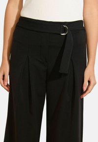 Čierne nohavice so širokými nohavicami, sklady vpredu a látkovým opaskom so striebornou D-krúžkovou prackou. Hladká textúra a priliehavý strih.