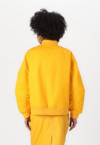 Giacca bomber gialla con colletto e polsini a costine, caratterizzata da una texture liscia e una vestibilità oversize. Include un piccolo etichetta del marchio sul retro.