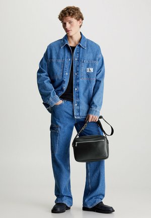 Giacca e pantaloni di denim di blu medio, con una patch del logo. Il modello tiene una borsa a tracolla in pelle nera con una cinghia decorata.