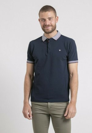 Homme aux cheveux courts et barbe, portant un polo bleu marine et un pantalon beige, debout devant un fond blanc uni.