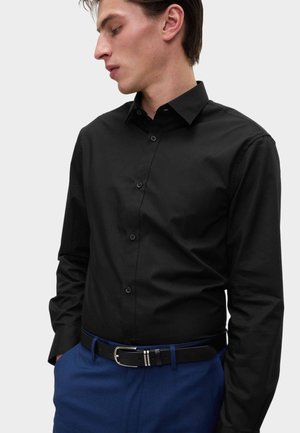 Camisa negra de botones con cuello puntiagudo, diseño ajustado y mangas largas. Combinada con un cinturón negro y pantalones azules.