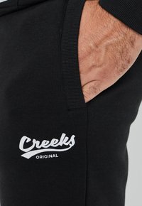 Zwarte joggingbroek met een wit "Creeks Original" logo, gemaakt van zachte stof met een ribgebreide tailleband en zijzakken.