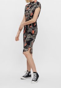 Abito nero a maniche corte con motivo di foglie tropicali e fiori in verde e arancione; presenta una vita legata e uno spacco laterale per la libertà di movimento.
