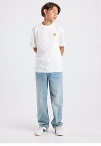 White short-sleeve t-shirt with a logo detail on the left chest, paired with light blue baggy jeans and white sneakers.