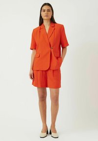 Oranssi, teksturoitu bleiseri lyhyillä hihoilla ja etunapilla, yhdistettynä vastaaviin shortseihin. Sisältää sivutaskut ja rakenteellisen muodon.