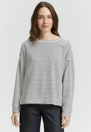 Kvinna med axellångt brunt hår iförd en vit långärmad skjorta med tunna svarta horisontella ränder och mörka jeans, stående mot en enfärgad bakgrund.
