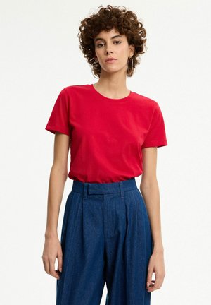 Mujer con cabello rizado que lleva una camisa roja de manga corta metida en unos pantalones azules de pierna ancha y cintura alta, de pie contra un fondo liso.