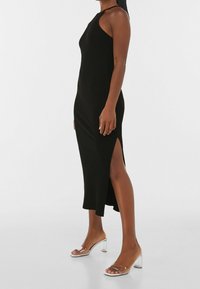 Robe midi noire côtelée sans manches avec une fente latérale. Texture lisse, coupe ajustée et style minimaliste complétés par des sandales à talons transparentes.