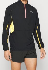 Giacca sportiva nera con pannelli gialli e una cerniera rosa, caratterizzata da un collo alto e polsini elasticizzati. Tessuto testurizzato per traspirabilità.