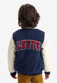 Námorní modrá a krémová varsity bunda s bílými rukávy, na zadní straně s červenobílou vyšívanou logem "LEVI'S". Měkká textilie.