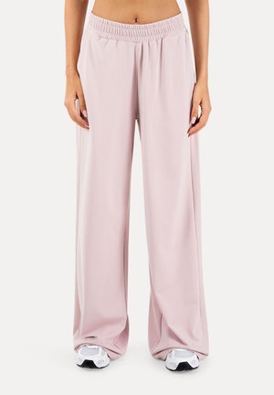 Frau trägt hellrosa weite Hosen mit elastischem Bund und weißen Sneakers, stehend vor einem einfarbigen Hintergrund.