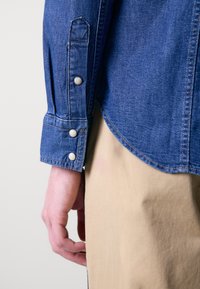 Denim-paita, jossa on sininen väri, napitettu hihansuu kolmella hopeapainikkeella ja kaareva helma. Yhdistetty vaaleanruskeisiin kangashousuihin.