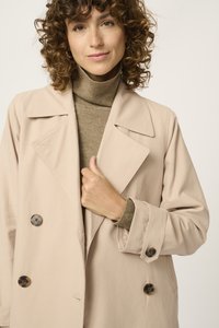 Abrigo beige de doble botonadura con grandes botones, ajuste ligeramente oversize y grandes solapas, superpuesto a un cuello alto ajustado marrón.
