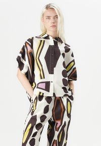 Chemise à manches courtes en tissu léger, présentant un motif abstrait audacieux en marron, jaune, rose et orange, avec un col et un bouton sur le devant.