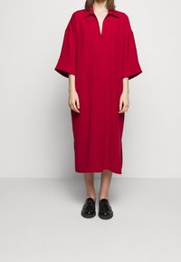 Robe rouge longueur genou avec col et manches courtes ; caractérisée par un décolleté en V et des poches latérales ; tissu lisse et coupe décontractée.